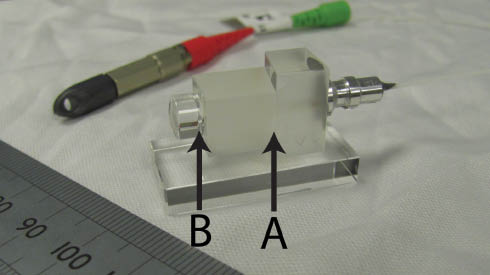 Figure 4 Photograph of a bonded fibre injector with beam expansion in glass, the ruler shows a scale in mm. The input AVIM connector can be seen in the background. The fibre terminates at bond interface A, and the collimating lens is bonded to a precision glass spacer at interface B. The fibre injector is bonded to a glass baseplate for mounting purposes. Reproduced from [56].
